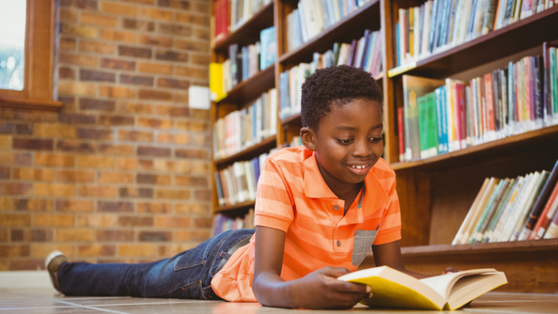 African American boy laying on ground in library reading a book - popular kids books