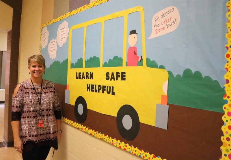 A woman stands beside a bulletin board of a yellow school bus with black words on it that say learn, safe, and helpful. There are bubbles with headlines like be respectful and bulleted items underneath.
