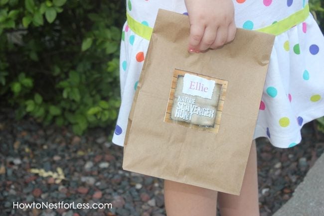 Child holding a paper bag labeled "Ellie: Nature Scavenger Hunt"