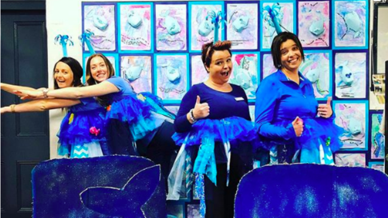 Four teachers wearing blue whale costumes in a classroom behind a cardboard cutout of a whale