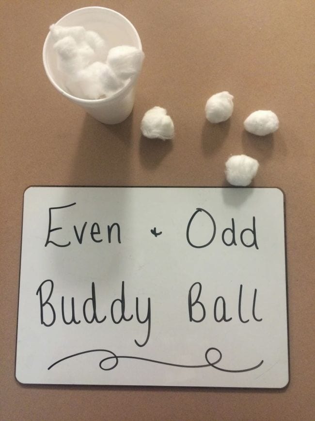 Whiteboard reading Even + Odd Buddy Ball next to a cup of cotton balls and four loose cotton balls