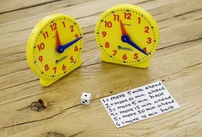 Two plastic toy clocks with a die and card with instructions for playing a second grade math game to learn to tell time