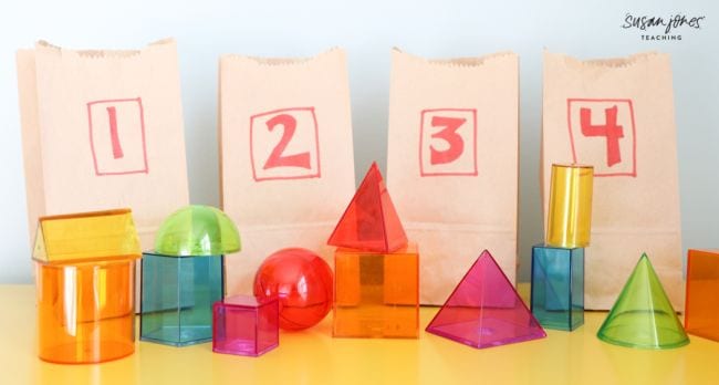 Colorful transparent 3-D shape blocks in front of paper bags numbered one through four (Second Grade Math Games)