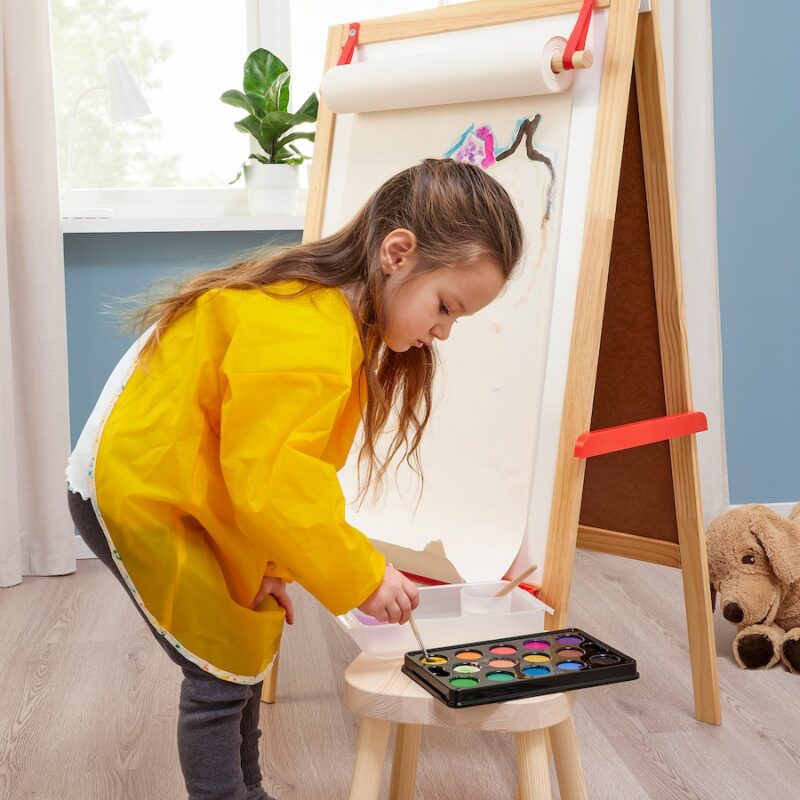 A little girl in a yellow smock stands before an easel painting.
