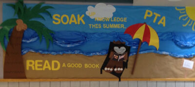 Bulletin board that reads 'Soak up knowledge this summer... read a good book.' An animal in a beach chair is reading a book. Water and sand are in the background with a palm tree in the corner. 