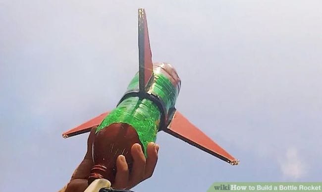 hand pointing a DIY rocket made from a green plastic bottle with brown cardboard wings up to the sky