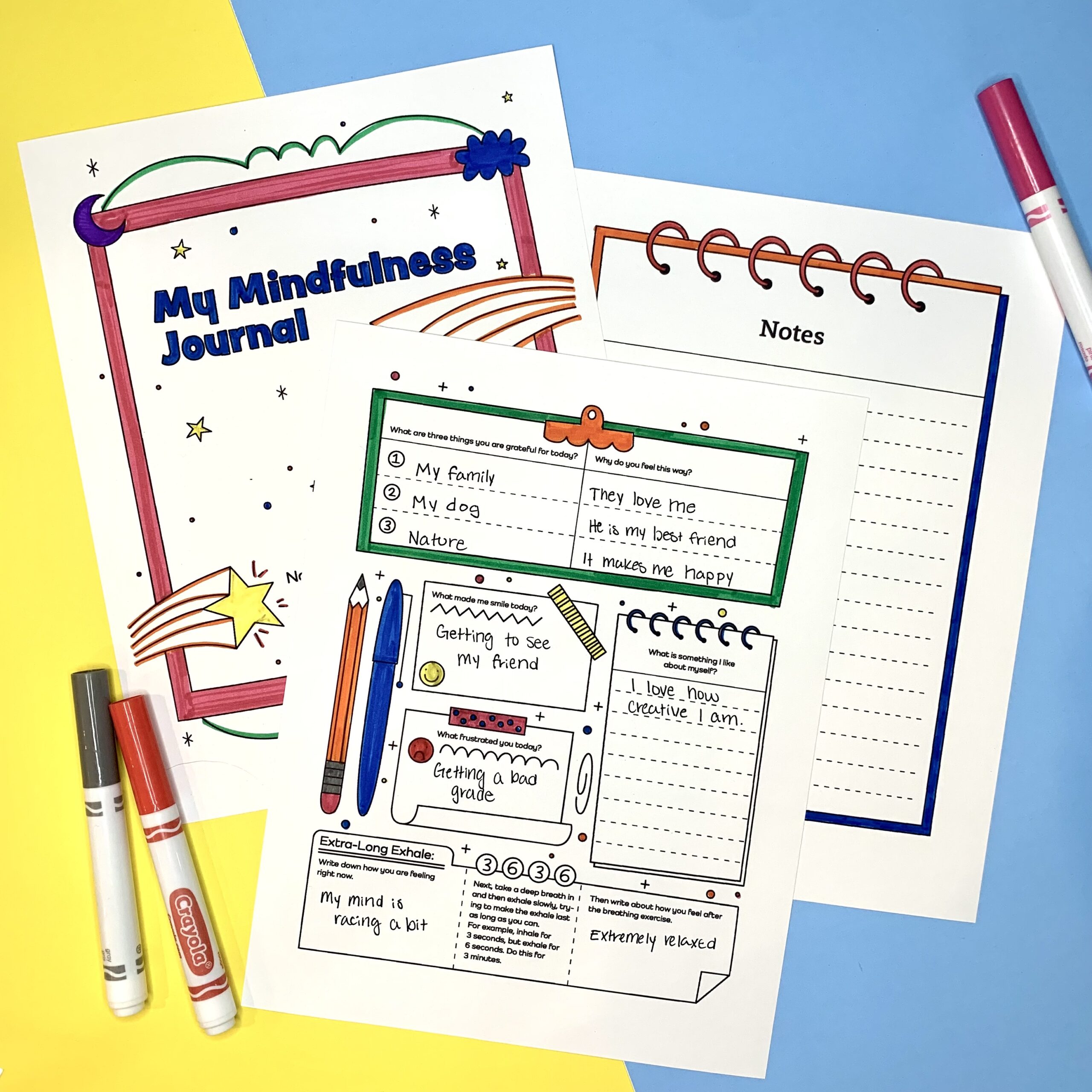 Photo of three pages from a mindfulness journal filled out with marker on a blue and yellow background with several markers lying around the pages