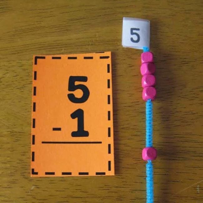 blue pipe cleaner with pink beads attached next to an orange card with a subtraction problem written on it