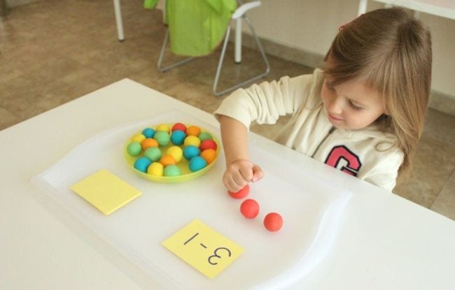 young girl at a table with balls of playdough in a dish and math problems on cards