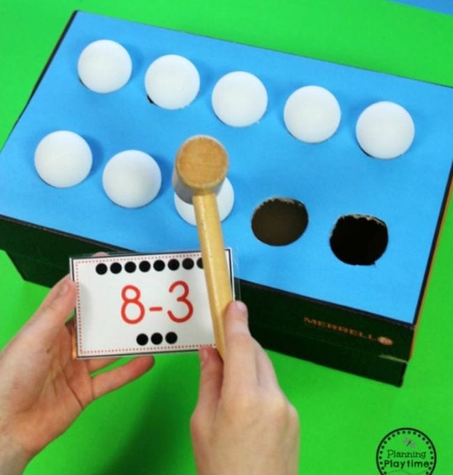 A colored paper-covered shoe box with holes cut on the top. In each of the holes is a ping pong ball. A hand holds a mallet and a math problem