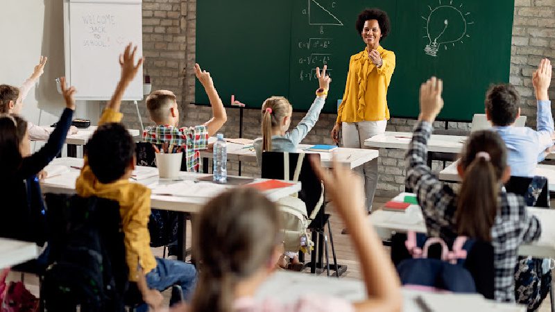 Happy African American teacher in front of a class and gesturing at her students who are raising their hands to answer the question in the classroom.