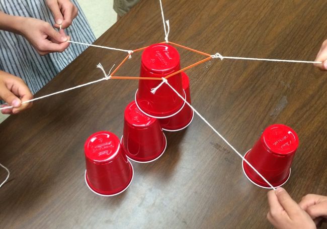 Group of people using string attached to a rubber band to grab and stack red plastic cups (Team Building Games for Adults)