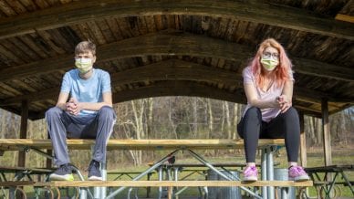 Teenage siblings wearing face masks sitting at a park grove outdoors with their face masks on keeping a social distance during the coronavirus pandemic.