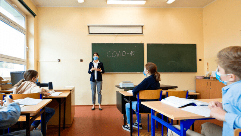 A teacher in her classroom with the words, "COVID-19" on the chalkboard. All students and the teacher are wearing masks.