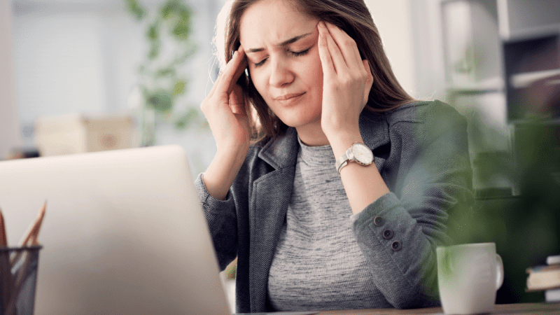 Brunette woman looking at a laptop while rubbing her temples due to a headache caused by working in a school with unhealthy air quality