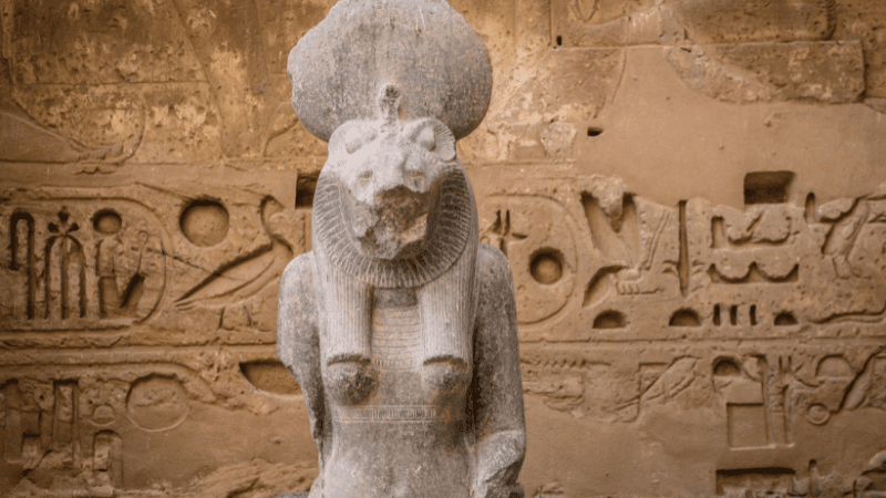 Statue of the Egyptian goddess Sekhmet in front of a stone wall covered in hieroglyphics 