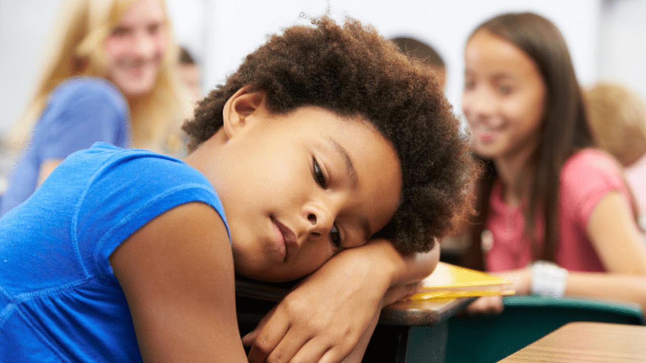 student with head down on desk and other students laughing in background - difference between meanness and bullying