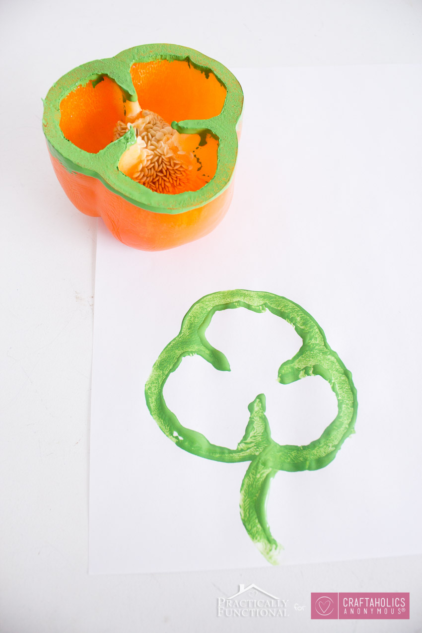 an orange bell pepper rimmed with green paint and a print of a shamrock on a piece of white paper, as an example of St. Patrick's Day activities 