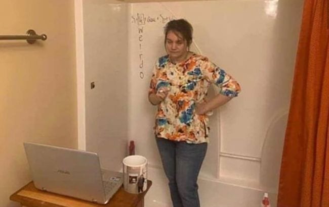 Teacher standing in a shower/bath stall doing virtual instruction writing with dry erase marker on the shower wall while in front of a laptop