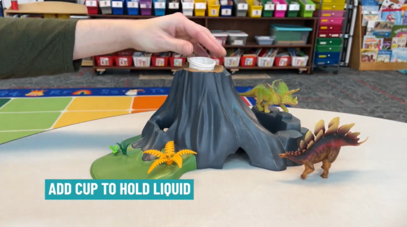 A hand is seen placing a paper cup in the top of a volcano mold. Text reads Add cup to hold liquid.
