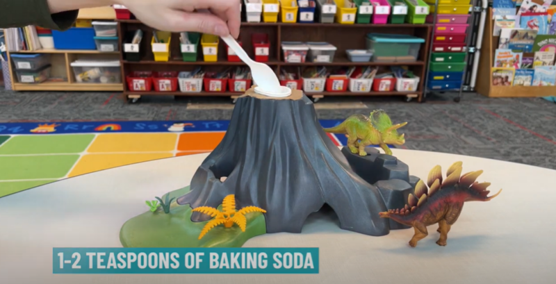 This step of a baking soda volcano shows a hand placing baking soda into a cup at the top of a volcano using a plastic spoon. Text reads 1-2 teaspoons of baking soda.