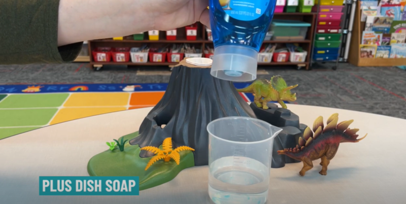 Text reads Plus Dish Soap. This step of a baking soda volcano shows dish soap being poured into a plastic measuring cup.