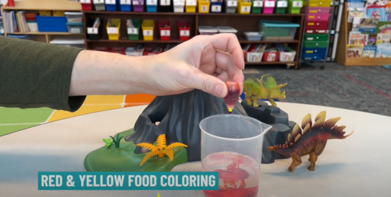 Text reads "red and yellow food coloring." A hand is seen squeezing a small food coloring container into a plastic measuring cup in this step of a baking soda volcano.