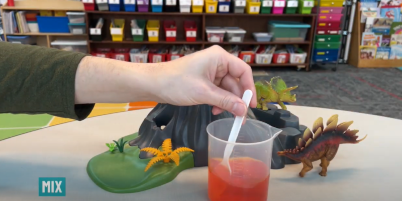 Text reads mix and a hand is seen mixing red liquid in a plastic measuring cup with a spoon.