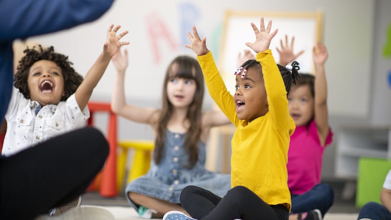 A multi-ethnic group of preschool students is sitting with their legs crossed on the floor in their classroom while raising their hands in the air.