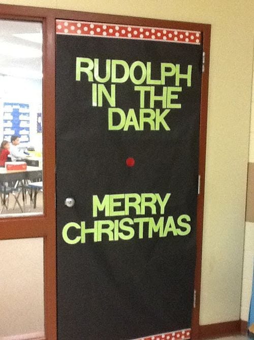 Winter classroom doors include this one that is black and has writing that says Rudolph in the Dark. There is a red nose on the black.