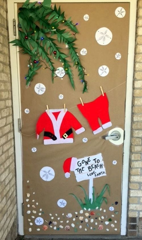 Classroom door decorated with a Santa outfit hanging on a line, palm trees made of paper, and seashells. Text reads "Gone to the Beach, Love Santa"