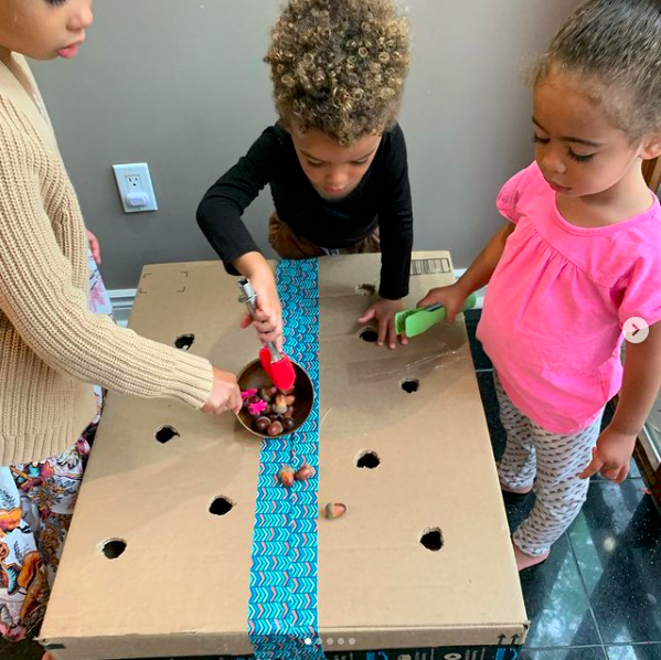 Three children using tweezers to drop acorns through a cardboard box with small holes cut in it