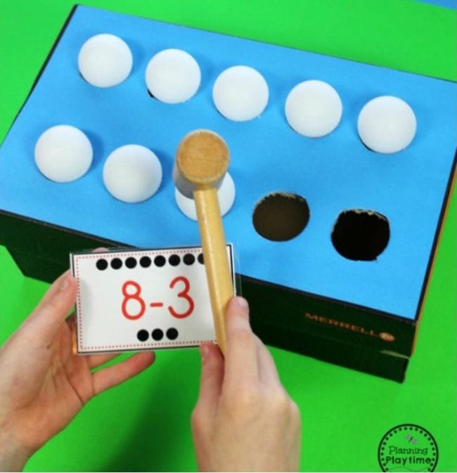 Student's hand holding a wood mallet and card saying 8 minus 3 over a box with ping pong balls resting in holes