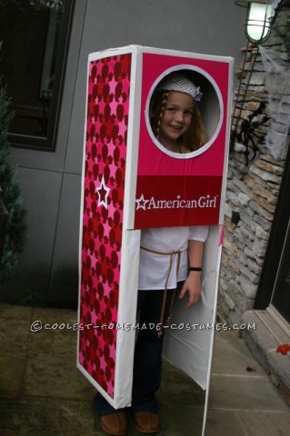 A girl is standing inside a pink and white box with her head showing through a cut-out. The American Girl logo is on the box.