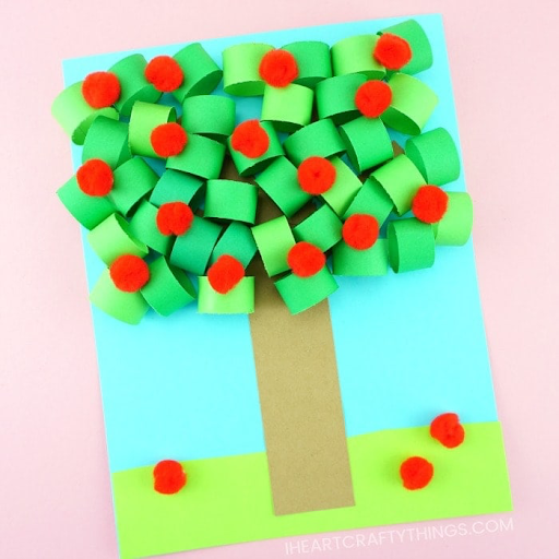 Apple tree craft made out of green construction paper rings, red pom poms, and brown paper for the trunk with a blue sky background and green grass.