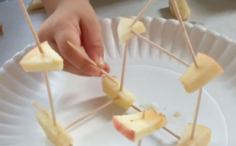 Child using toothpicks and pieces of apple to build a tower on a paper plate