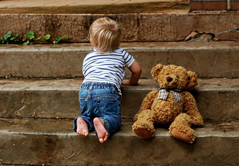 Baby crawling up stairs outside with a teddy bear sitting on stairs.