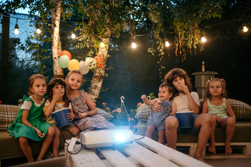 Group of children are sitting in the yard during a birthday party and watching a movie on a video projector