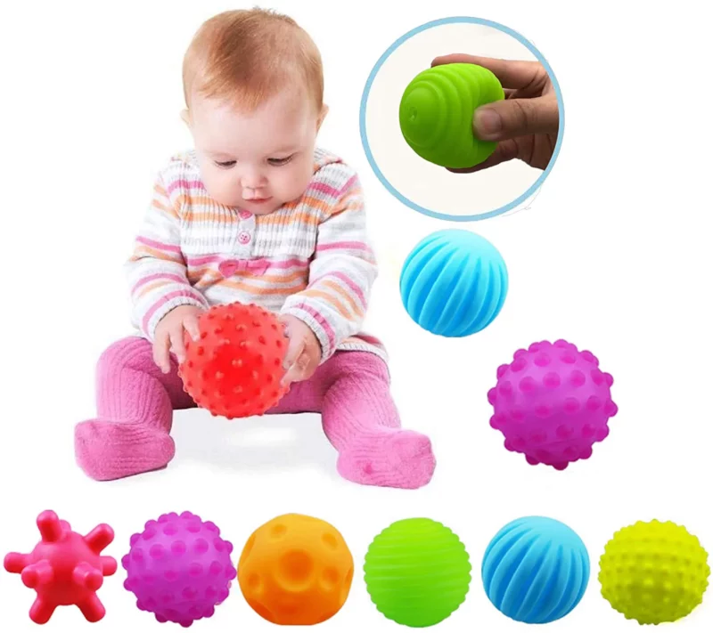 A baby sits playing with a red ball. Other balls in different colors and shapes are lined up in front of her.