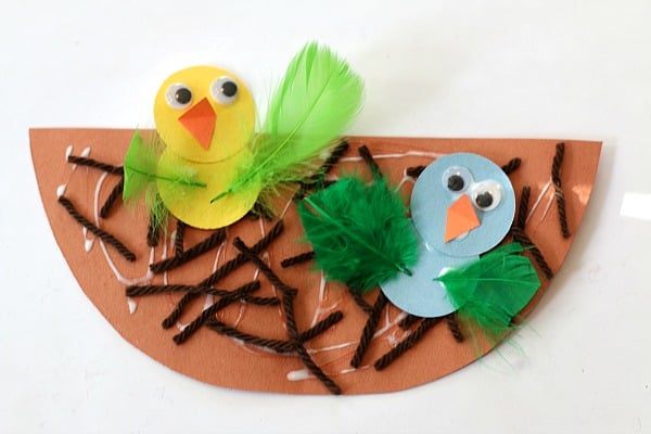 Two baby birds made from construction paper and feathers sit atop a nest as an example of summer crafts for kids 