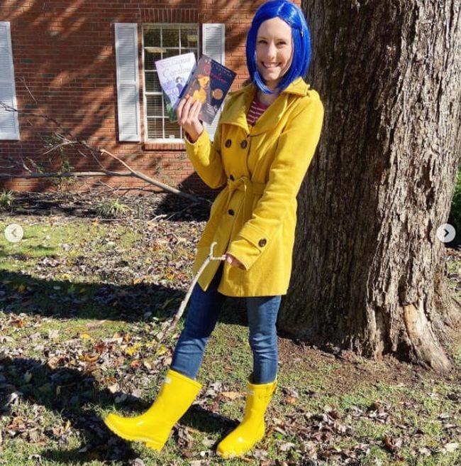 A woman stands in yellow rainboots, a yellow trenchcoat, and a blue wig, holding a key necklace.