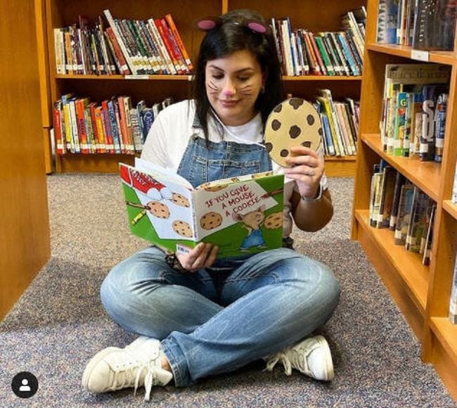 Woman in blue overalls and mouse ears holding a large paper cookie and reading a book (Book Character Costume Ideas)