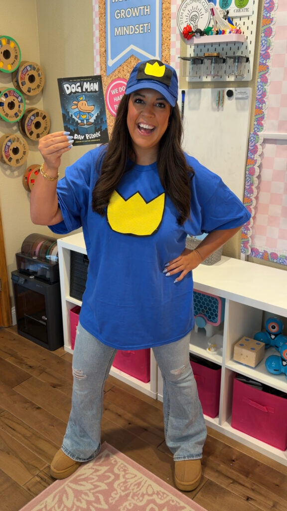 A teacher poses in her classroom wearing a t-short and hat from the Dogman book series. She is also holding a copy of the book.
