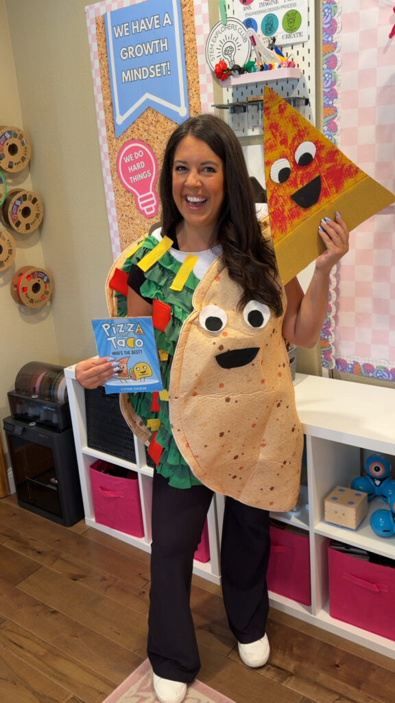 A teacher poses in her classroom dressed as a taco and holding a cardboard piece of pizza, along with the book Pizza & Taco