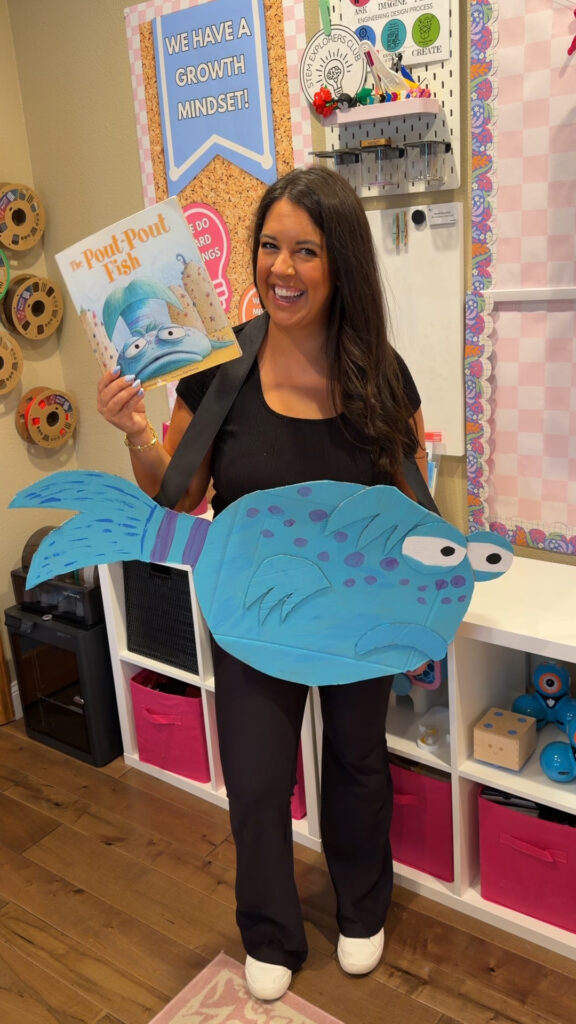 A teacher poses in her classroom wearing a cardboard Pout Pout Fish costume and holding the book