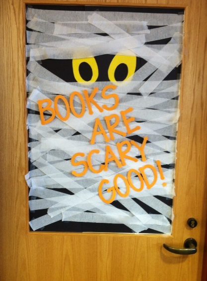 A door is decorated to show two eyes peeking out from white crepe paper designed to look like a mummy. Text in orange reads books are scary good.