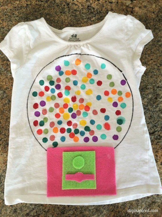 A white t-shirt has a bubble gum machine on it. There are 100 painted on dots for the bubble gum and some felt has been used to make the dispenser part of the machine (100th day of school shirt ideas)