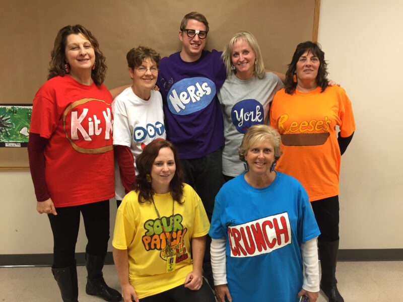 Seven teachers pose wearing t-shirts with different candy logos on them.