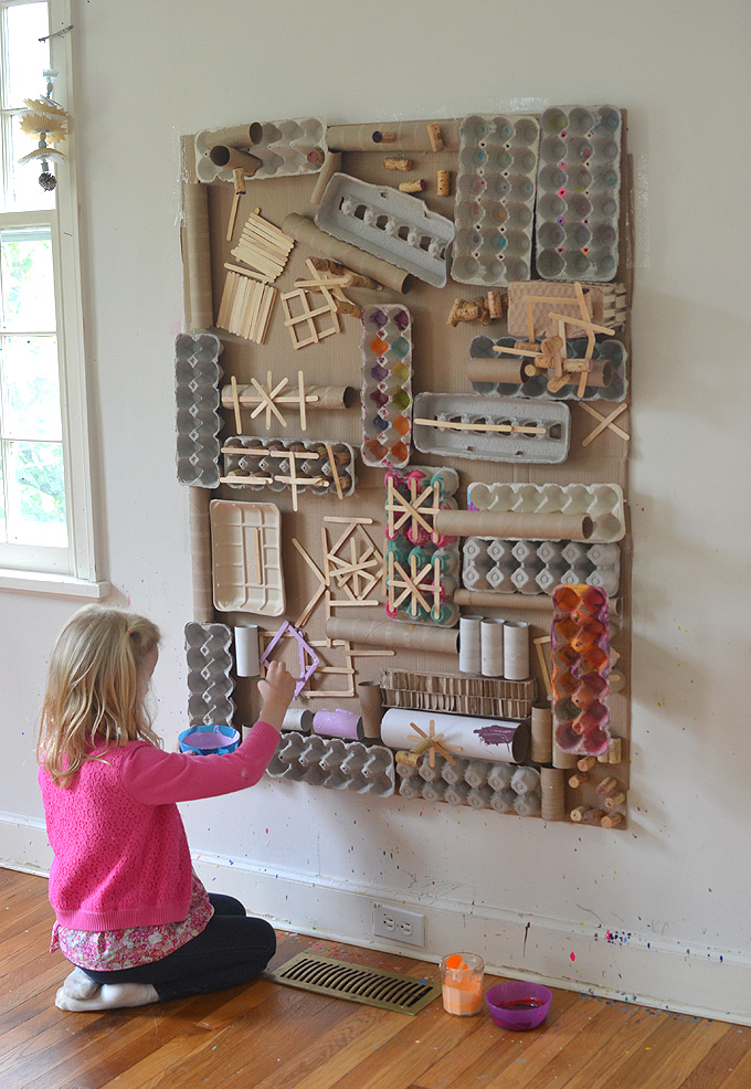A large display of recycled cardboard containers, etc. is shown attached to a wall. A little girl sits in front of it attaching more to it.