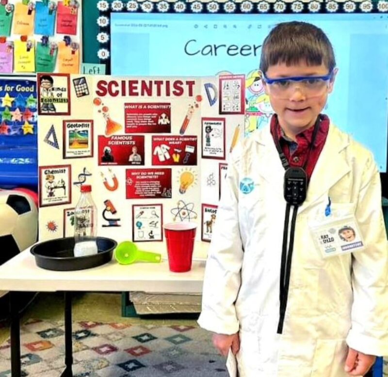 Elementary student dressed in a lab coat and goggles, with a poster about being a scientist